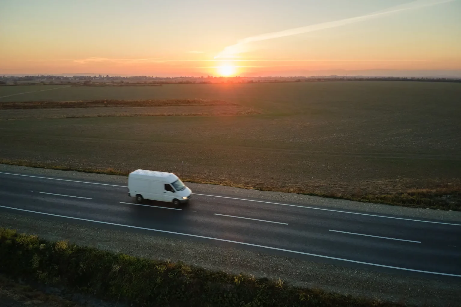 Un fourgon utilitaire blanc roulant sur une route de campagne au coucher du soleil, avec un paysage agricole et un ciel dramatique en arrière-plan.