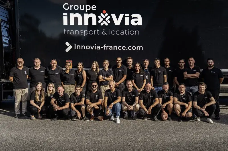 Un grand groupe de personnes portant des chemises noires avec le logo "Groupe innovia" est réuni pour une photo d'équipe. Ils se tiennent devant un camion semi-remorque portant la marque de l'entreprise. L'équipe est diversifiée, avec des membres d'âges et de sexes différents.