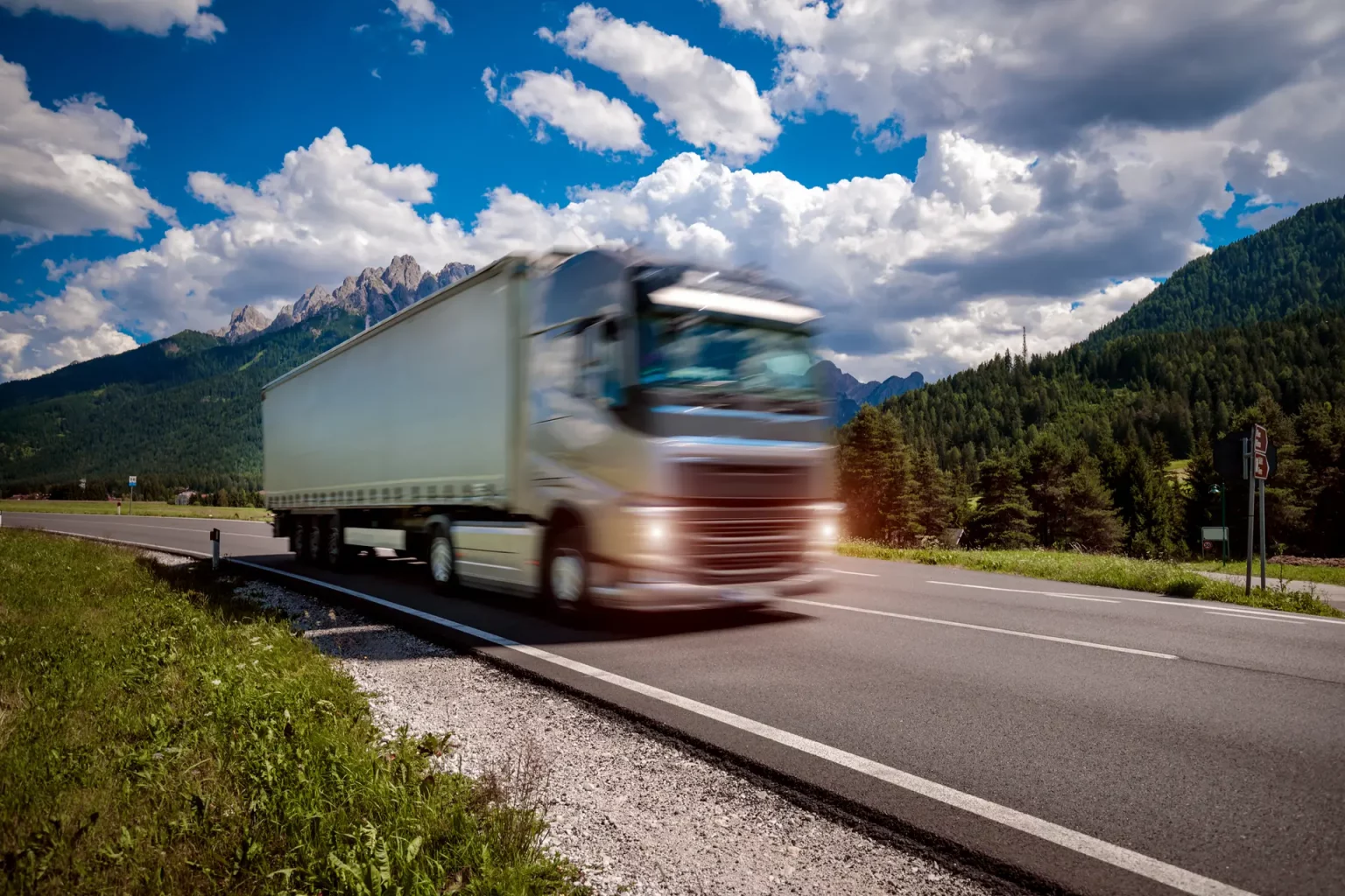 Camion de transport à semi-remorque roulant à grande vitesse sur une autoroute avec des montagnes et un ciel bleu nuageux en arrière-plan. L'effet de mouvement est capturé avec un flou de vitesse sur le camion et la route.