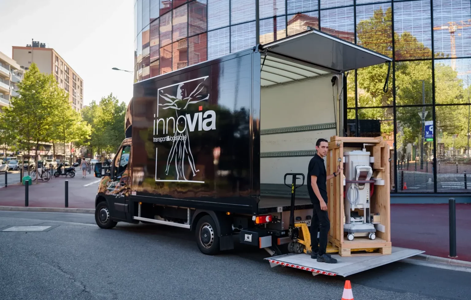 Une camionnette de livraison, dont les portes arrière sont ouvertes, est garée dans une rue de la ville. Un jeune homme utilise un transpalette pour charger une caisse en bois dans le véhicule. Des cônes de signalisation délimitent la zone autour de la camionnette afin d'assurer la sécurité pendant le processus de chargement.