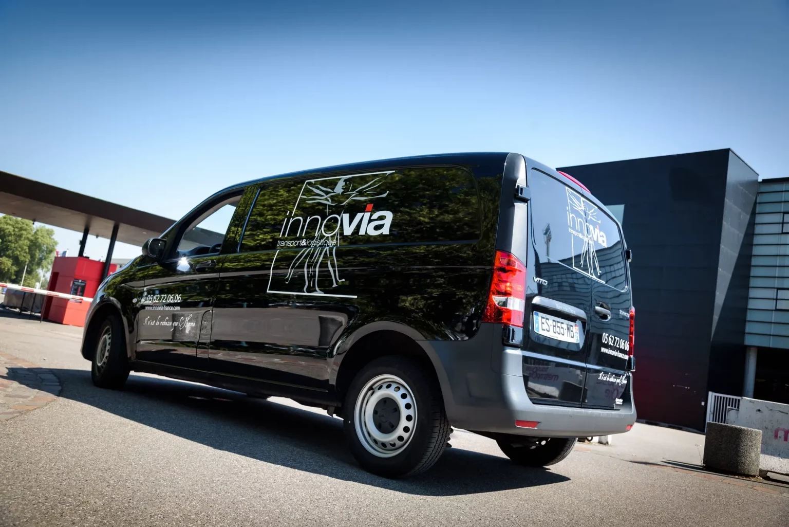 Garée sous un ciel bleu clair, la camionnette est bien visible. La marque "Groupe innovia" est visible sur les panneaux latéraux et arrière du véhicule. La camionnette est garée près d'un bâtiment moderne au design épuré.