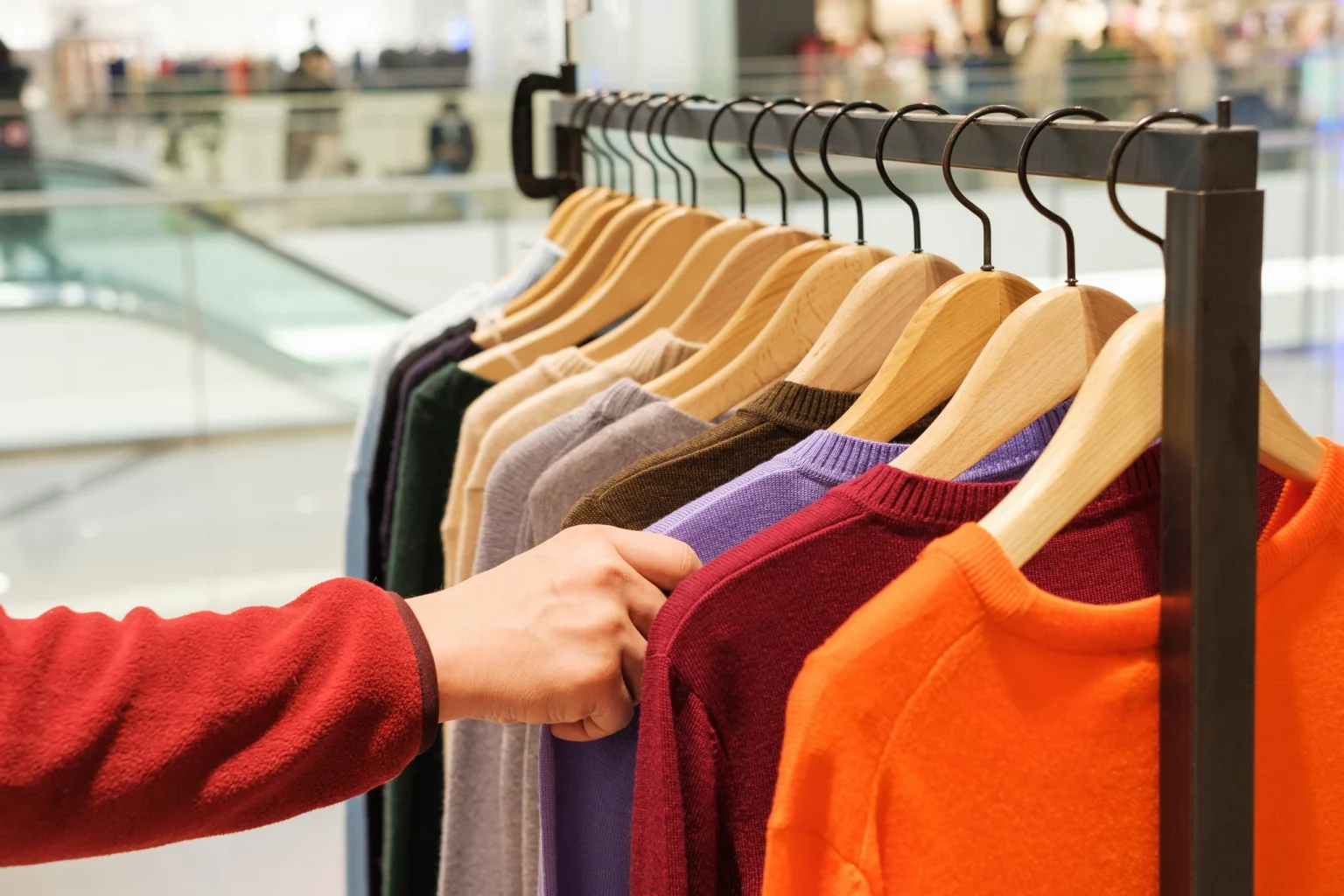 Une main sélectionnant un pull coloré parmi une gamme de vêtements sur des cintres dans un magasin de détail.