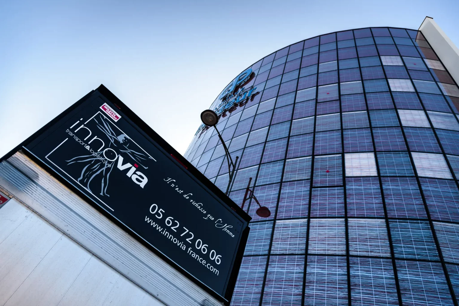 Un grand panneau d'affichage portant la marque "Groupe innovia" et ses coordonnées est monté sur le côté d'un immeuble de bureaux en verre. Le bâtiment reflète le ciel bleu et l'environnement sur sa surface réfléchissante.