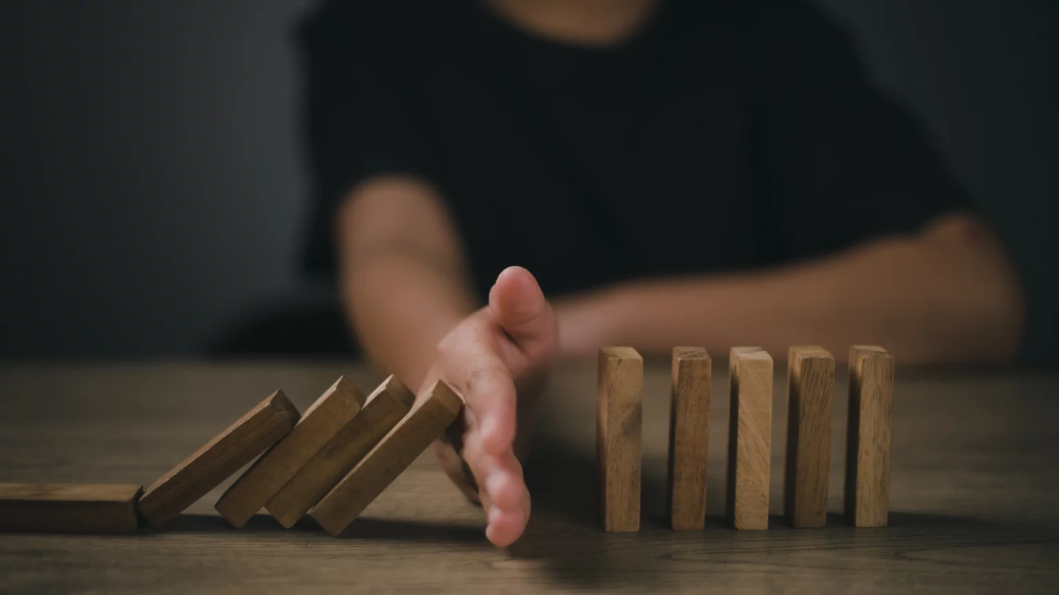 Une main d'adulte stoppant la chute de dominos en bois alignés sur une table, symbolisant la gestion du risque et la protection contre les effets en chaîne négatifs dans une ambiance sombre et sérieuse.