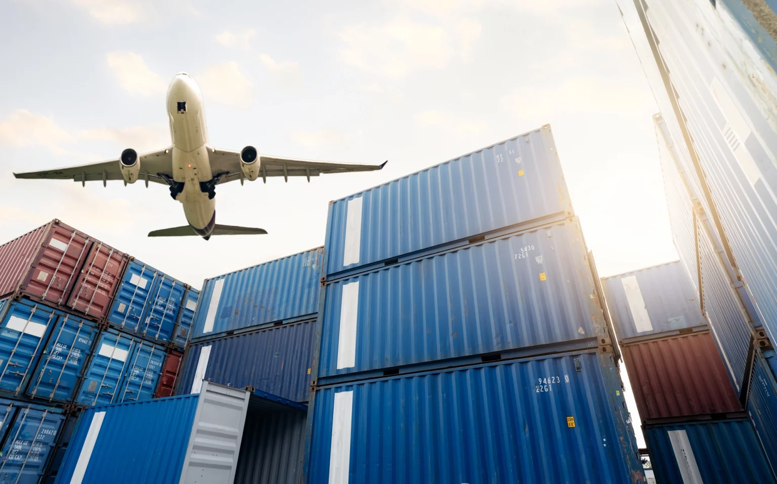 Avion-cargo en vol au-dessus d'une pile de conteneurs d'expédition, symbolisant les services de logistique aérienne pour le transport de marchandises à l'échelle mondiale.