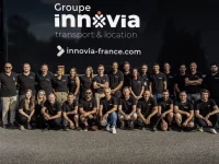 Un grand groupe de personnes portant des chemises noires avec le logo "Groupe innovia" est réuni pour une photo d'équipe. Ils se tiennent devant un camion semi-remorque portant la marque de l'entreprise. L'équipe est diversifiée, avec des membres d'âges et de sexes différents.